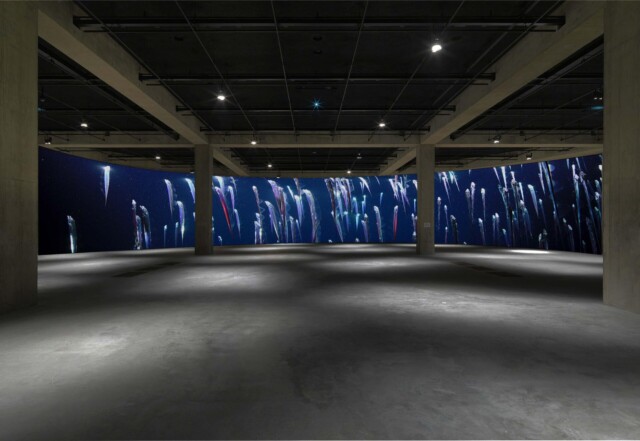Kuvassa näyttelytila, jonka takaseinälle on installoitu suuri sinisävyinen video- tai valokuvateos