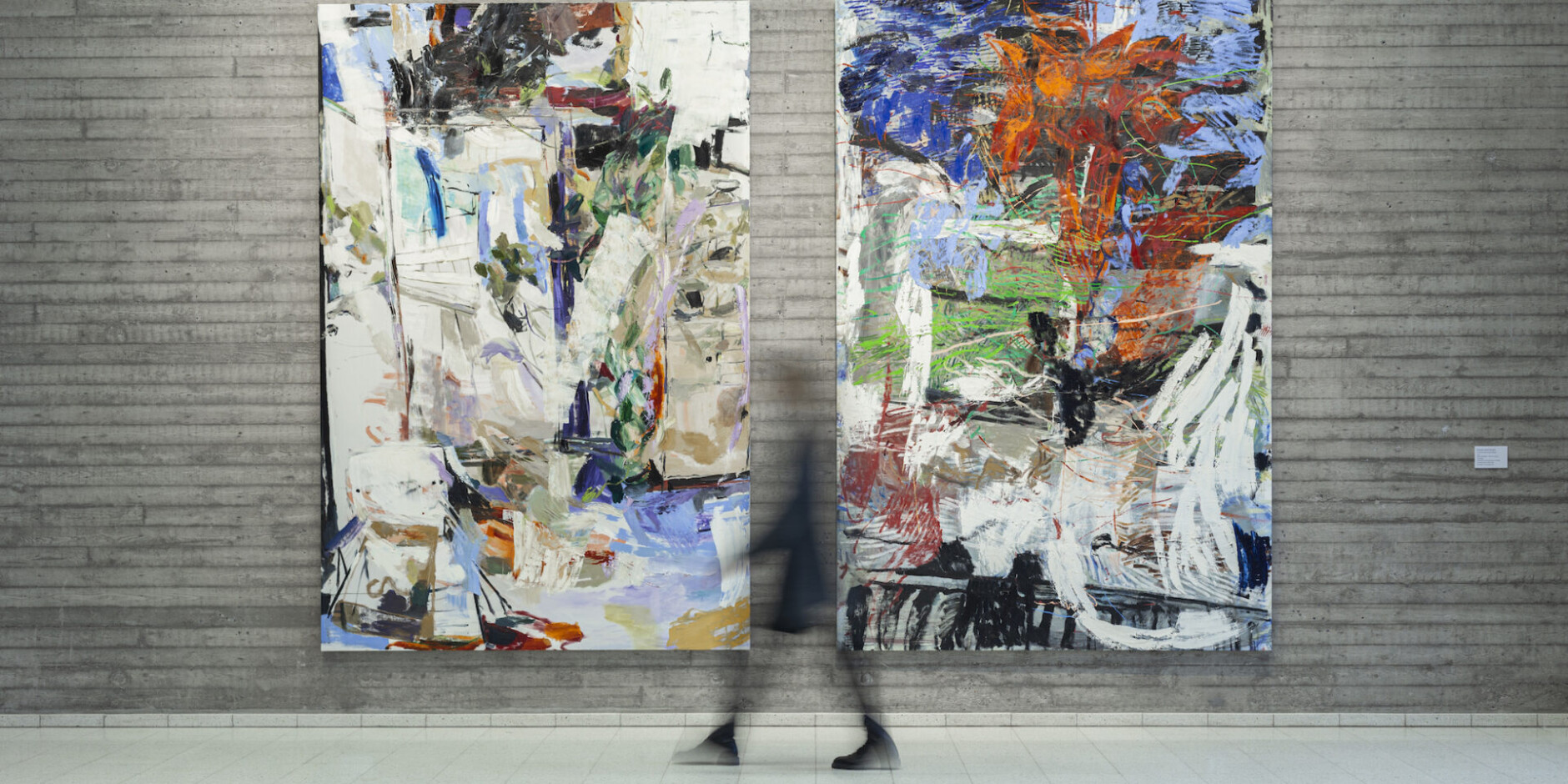 Taidemuseossa betoniseinällä roikkuu kaksi samankokoista värikästä, abstraktia Anna Retulaisen maalausta. Valokuvassa epätarkka ihmishahmo kävelee maalausten ohi.