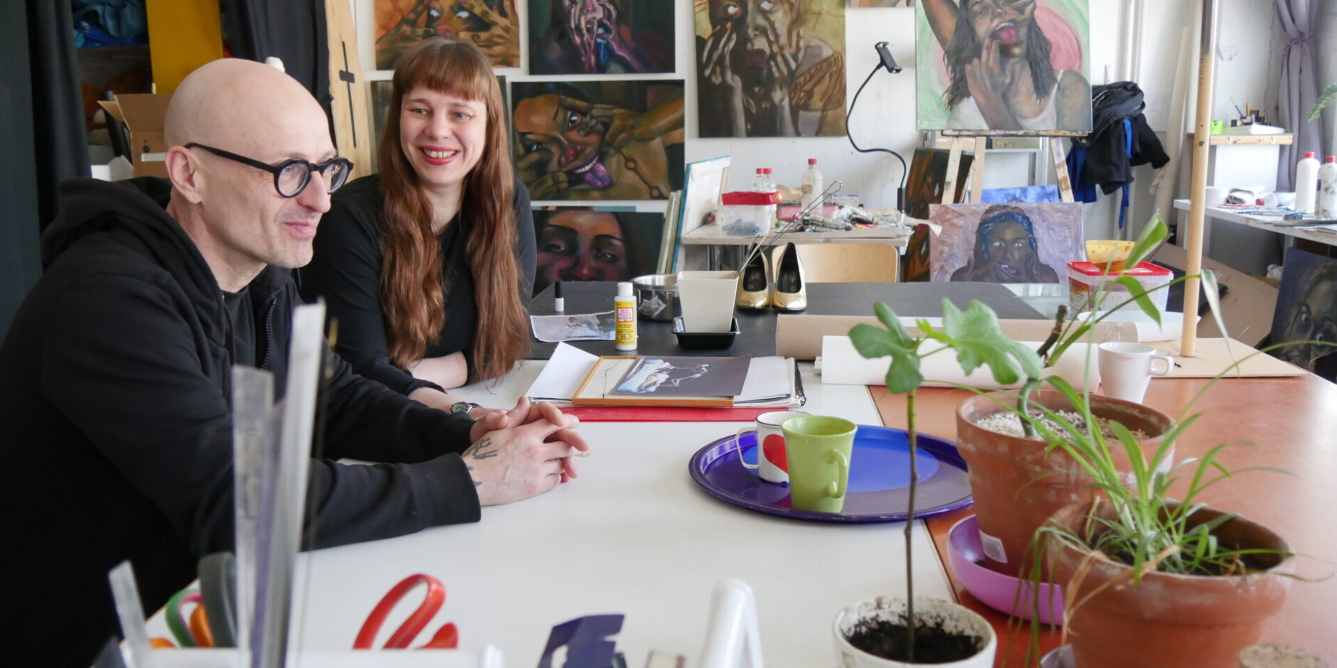 Taiteilijat Katri Kainulainen ja Maximilian Latva istuvat työhuoneellaan pöydän ääressä, taustalla maalauksia