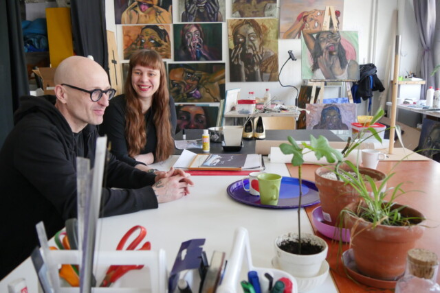 Taiteilijat Katri Kainulainen ja Maximilian Latva istuvat työhuoneellaan pöydän ääressä, taustalla maalauksia